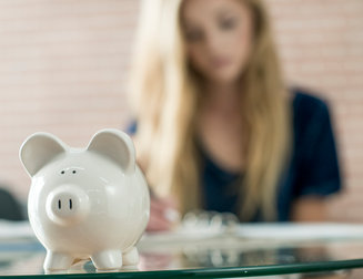 A high school teenager is writing down monthly budget in her notebook. A piggy bank is sitting on the table.